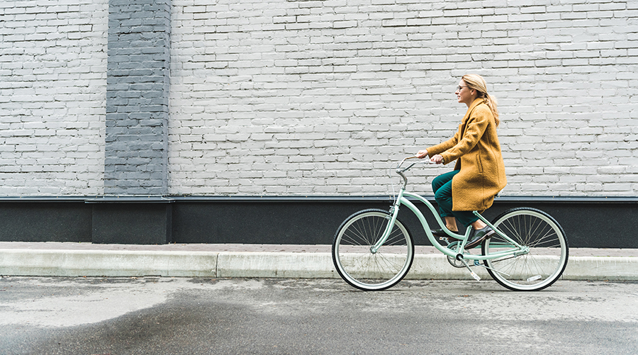 Kuvituskuva, jossa keltatakkinen nainen ajaa polkupyörällä valkoisen tiiliseinä edessä.